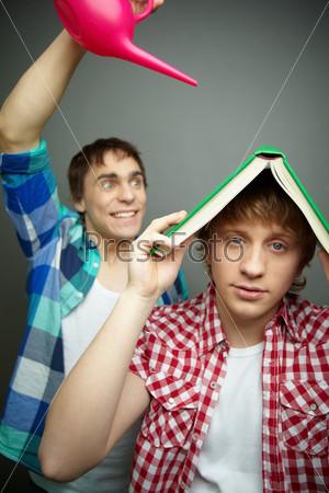 Cheerful guy pouring water from a pot while his friend using book as shelter, fool\'s day series