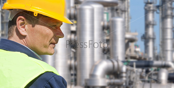 Close up of an engineer wearing a safety vest, blue coveralls, and a hard hat in front of a petrochemical plant