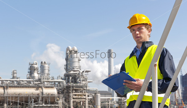 Young supervisor with a note board and pen in his hands, wearing a hard hat and safety vest in front of a petrochemical plant and refinery