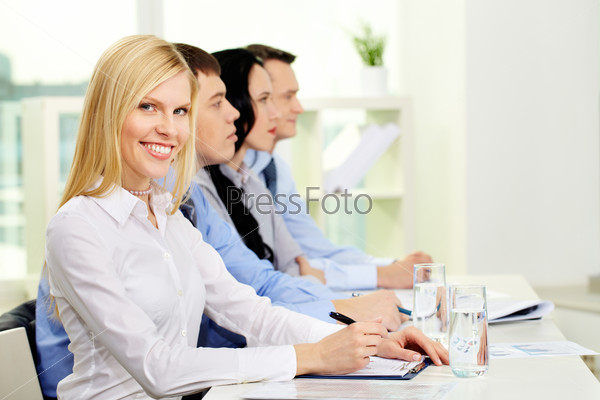 Pretty business lady looking at camera while her colleagues listening to someone off-screen