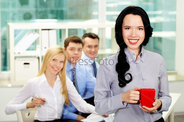 Cheerful lady holding a cup during coffee break, her colleagues smiling from behind her back