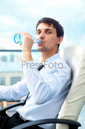 Young business man drinking water sitting relaxed on armchair at his modern office