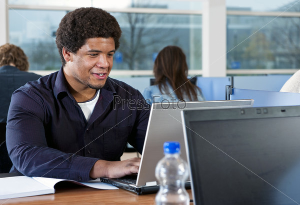 Young African man, studying in a library behind a laptop and his college notes with more people in the background