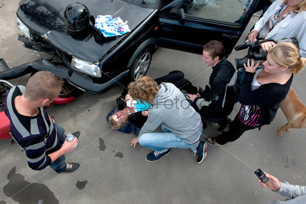 Chaotic scene just after a car crash, with an injured woman lying on the ground, several bystanders providing first aid, and a television reporter with camera capturing the scene, seen from above