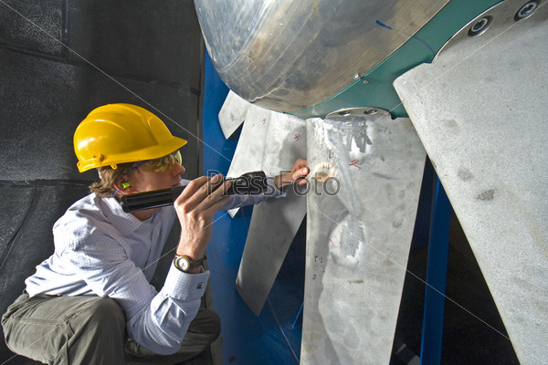 A young engineer inspecting the blades of an industrial wind tunnel wearing a hard top, earplugs and protective goggles