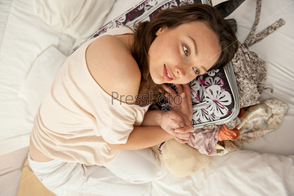 Beautiful young slim woman on the bed indoors unpacking clothes from suitcase