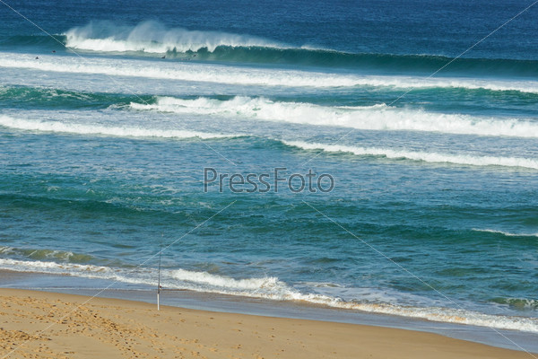 Swell and fishing rod set up at sandy beach while large waves breaks with spray and surfers diving under wave in Mornington Peninsula, Victoria, Australia