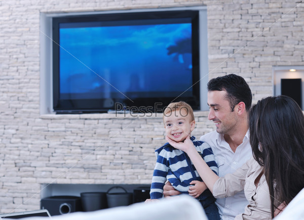 Счастливая молодая семья отдыхает в новой квартире на фоне современного телевизора