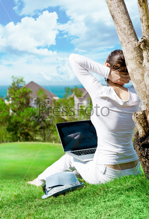 Young woman sitting under tree with laptop and dreaming. Idyllic outdoor scenery