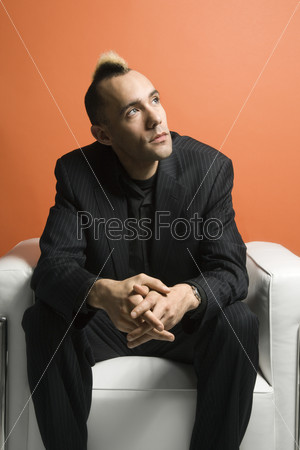 Caucasian man with mohawk wearing suit sitting in chair against orange background.