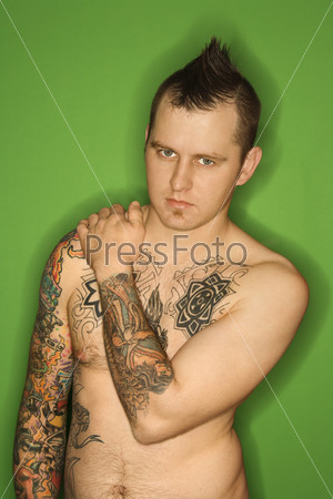 Caucasian man with mohawk and tattoos standing against green background.