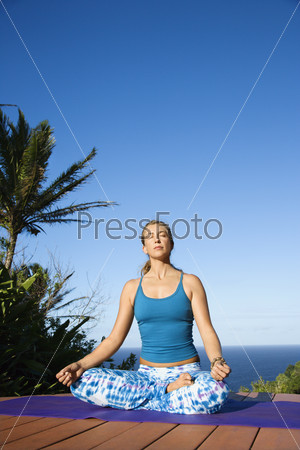 Attractive young woman in blue sits on an exercise mat doing yoga with the ocean in the background. Vertical shot.