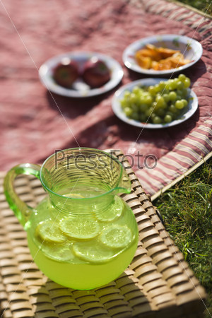 Green plastic pitcher of lemonade and lemons with a picnic spread out in the background.