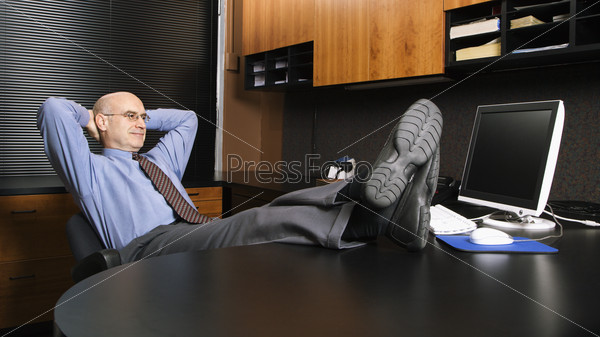 Caucasian middle-aged businessman in office sitting with feet on desk.