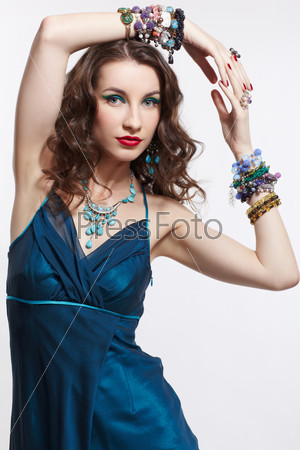 portrait of young beautiful brunette woman in blue dress, bracelets, rings, ear-rings and beads posing on gray