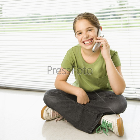Caucasian preteen girl sitting on floor talking on cell phone and smiling at viewer.