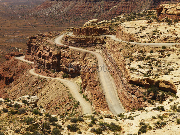 High angle view of a winding dirt road on a desert rock formation. The surrounding landscape is visible in the background. Horizontal shot.