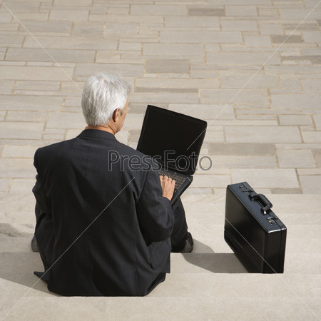 Back view of Caucasian middle aged businessman sitting on steps outdoors with laptop and briefcase.