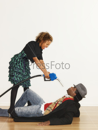 Man on floor with angry woman standing over him pointing gasoline pump nozzle at him like a gun.