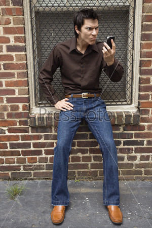 Confused young Asian man next to brick wall and window looking at cell phone.