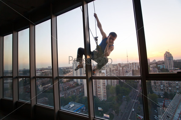 Industrial climber looking through a window while hanging on a rope outside a high building