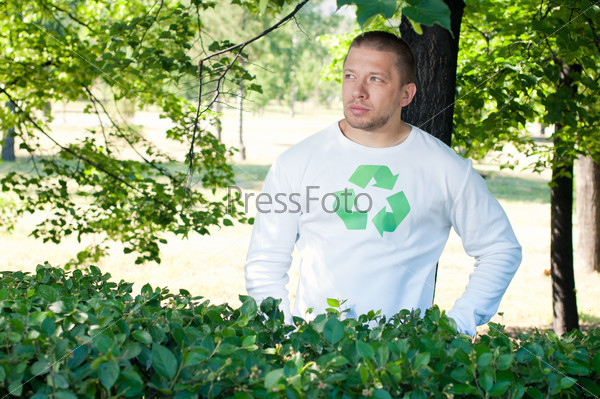 An adult man with recycle logo on his shirt standing under the green tree