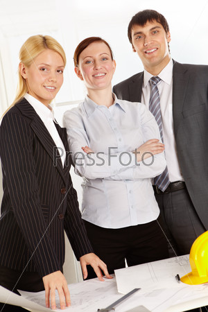 Three business people standing at table with plan, looking at camera and smiling