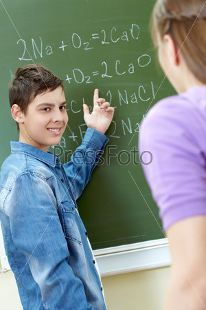 Clever boy pointing at blackboard while explaining formula to classmate