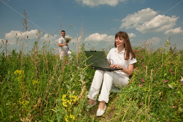 Happy Young Woman on the grass field with a laptop against the background of her beloved husband with a bouquet of flowers in their hands