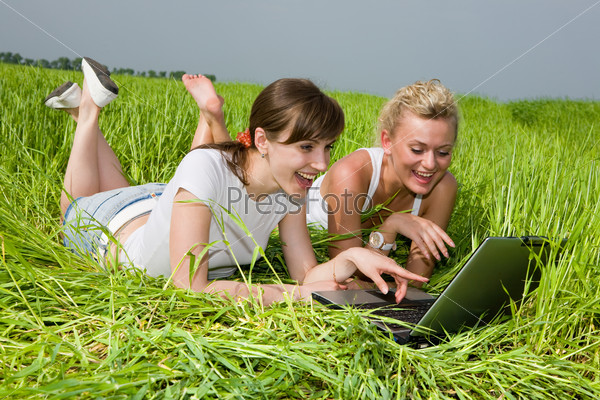 Two beautiful girls in white clothes are laughing near laptop computer outdoors. Lay on the green grass