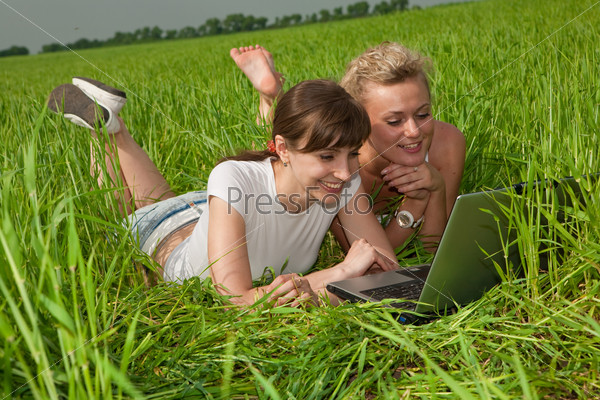 Two beautiful girls in white clothes are laughing and looking at laptop computer outdoors. Lay on the green grass.