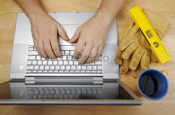 Contractor Reviews Project on Laptop with level, gloves and coffee to his side. Great image for online information regarding home improvement, additions, remodeling or construction.