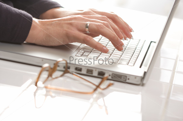 Female hands typing on the keyboard of the laptop with eyeglasses nearby.