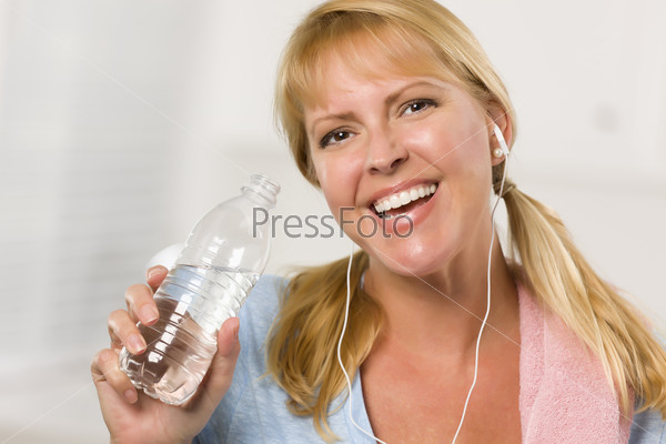 Pretty Blonde Woman with Towel and Ear Phones Drinking From Water Bottle in Her Kitchen.