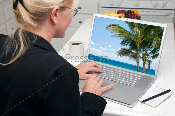 Woman In Kitchen Using Laptop to Research Travel and Vacations. Screen image can easily be replaced using the included clipping path.