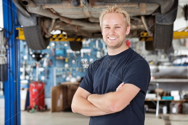 Portrait of cheerful young mechanic with arms crossed standing in his auto repair shop