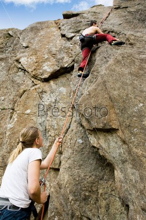 A female belaying a male on a steep rock face.  Shallow depth of field with the focus on the climbing which is belaying (the on at the bottom)