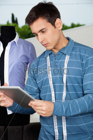 Young male tailor in casual t-shirt with measuring tape around neck holding tablet PC
