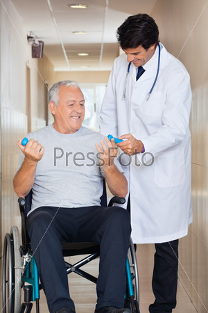 Happy young doctor giving hand weights to the senior man sitting in a wheelchair at hospital