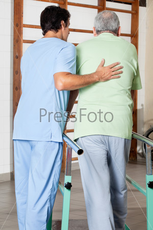 Rear view of physical therapist assisting senior man to walk with the support of bars at hospital gym