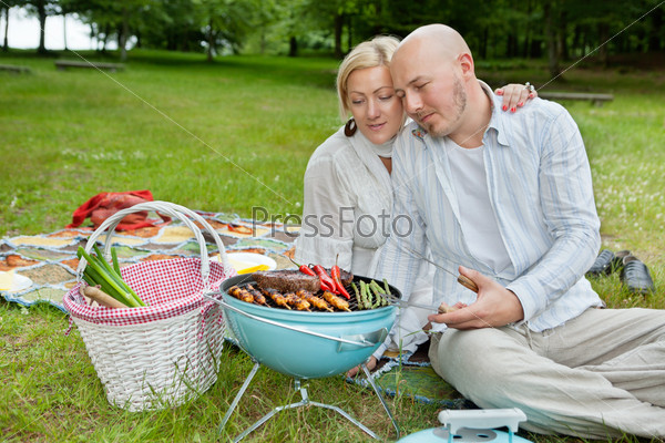 Mature couple in casual wear cooking meat and chilies on a portable barbecue at an outdoor picnic in forest park