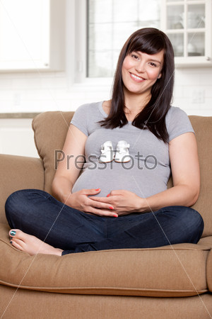 Portrait of expecting mother sitting on sofa with baby shoes on belly