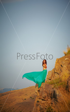 Beautiful girl standing on a precipice. Turquoise cape fluttering over the precipice