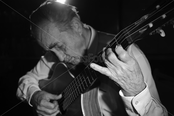 An elderly man in white shirt playing an acoustic guitar. Dark background. Monochrome.  Focus on the hand.