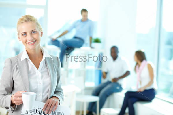 Portrait of happy female with cup and newspaper looking at camera in working environment, stock photo