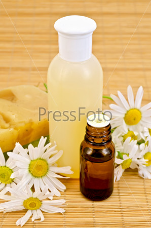 Oil in a bottle, lotion in a plastic bottle, two homemade soaps, chamomile flowers on a bamboo mat