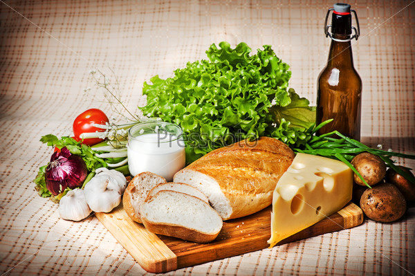 Помидоры, зелень, чеснок, картофель, лук и свежий хлеб, сыр и стакан молока на старой скатерти