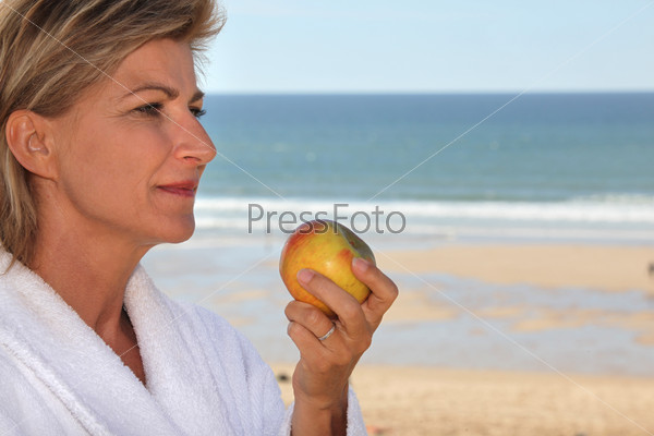 mature blonde woman dressed in bathrobe eating an apple in front of the sea