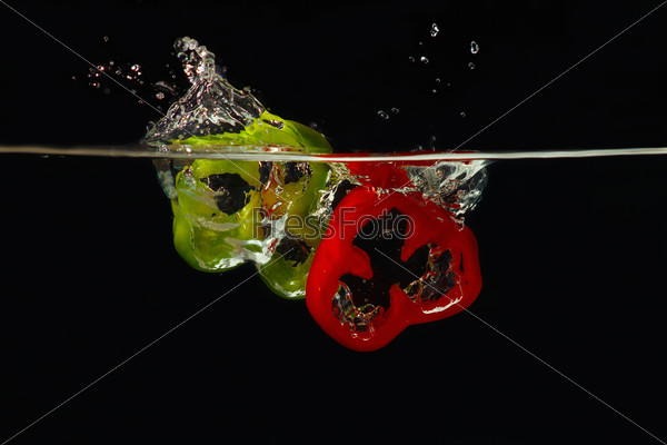 Slices of fresh red and green peppers falling into the water with a splash on black background