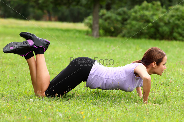 Young and cute sporty girl do her push-ups exercises in the park on a green lawn